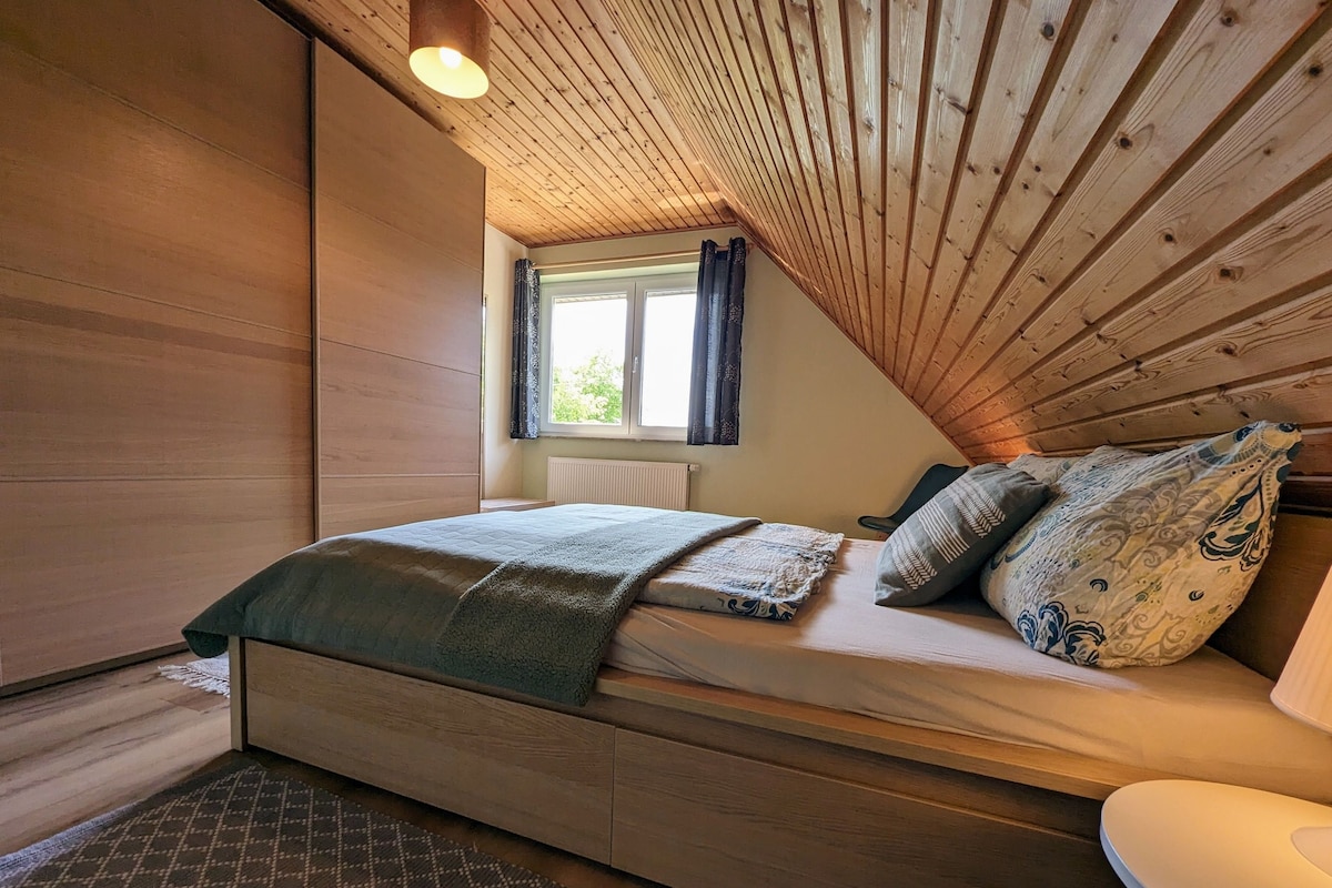 A cozy bedroom with a wooden ceiling, a bed with patterned bedding, a window with curtains, and a radiator.