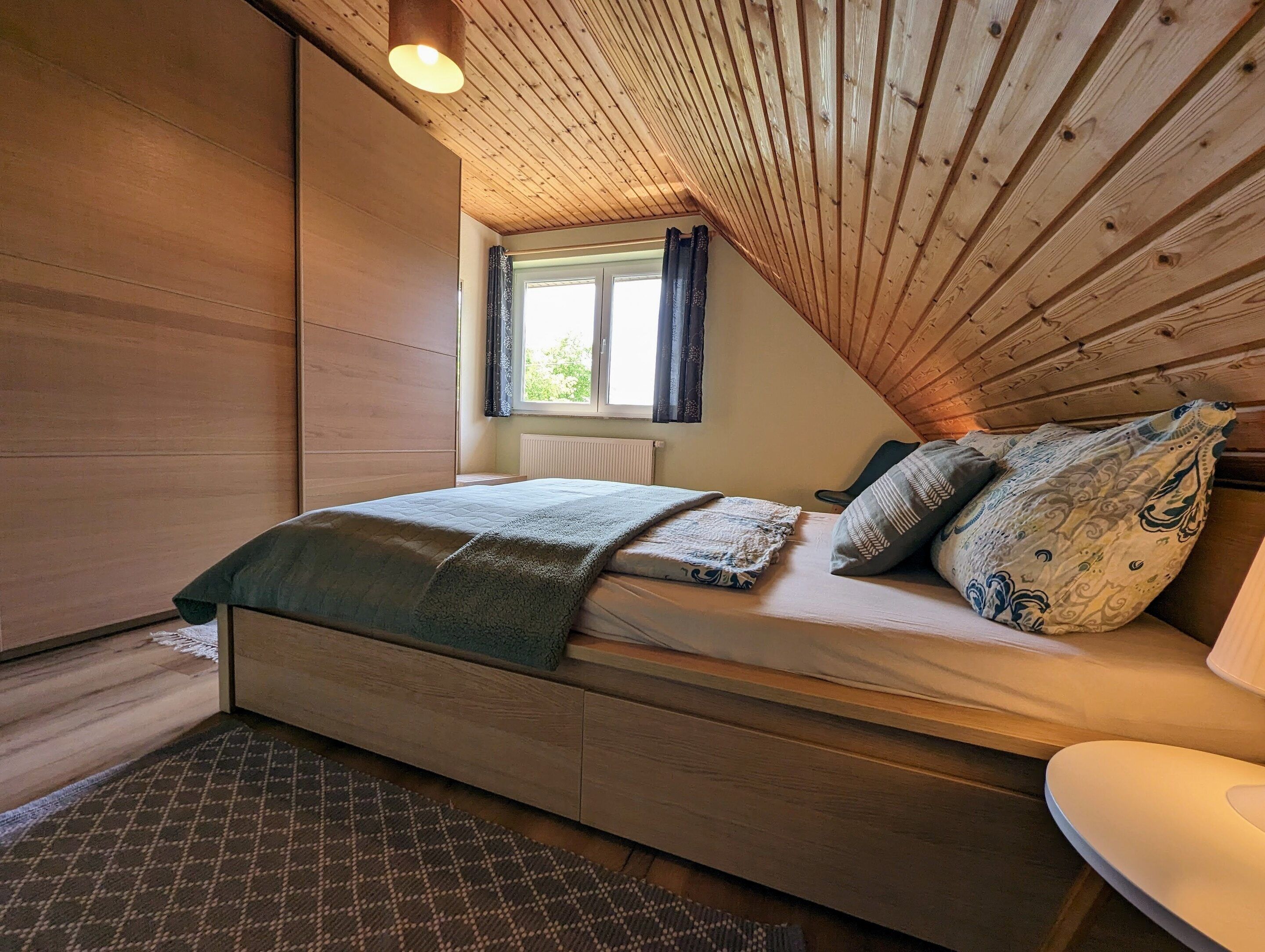 A cozy bedroom with a wooden ceiling, a bed with patterned bedding, a window with curtains, and a radiator.