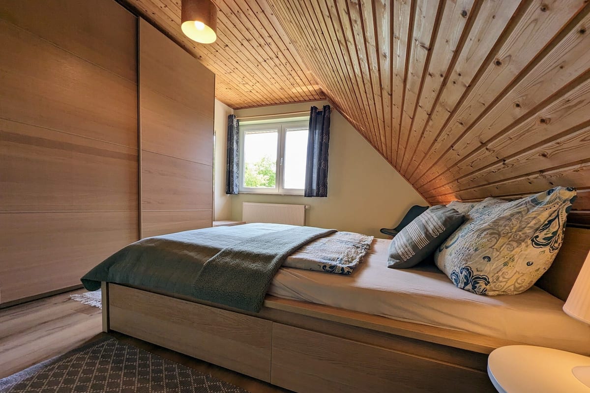 A cozy bedroom with a wooden ceiling, a bed with patterned bedding, a window with curtains, and a radiator.