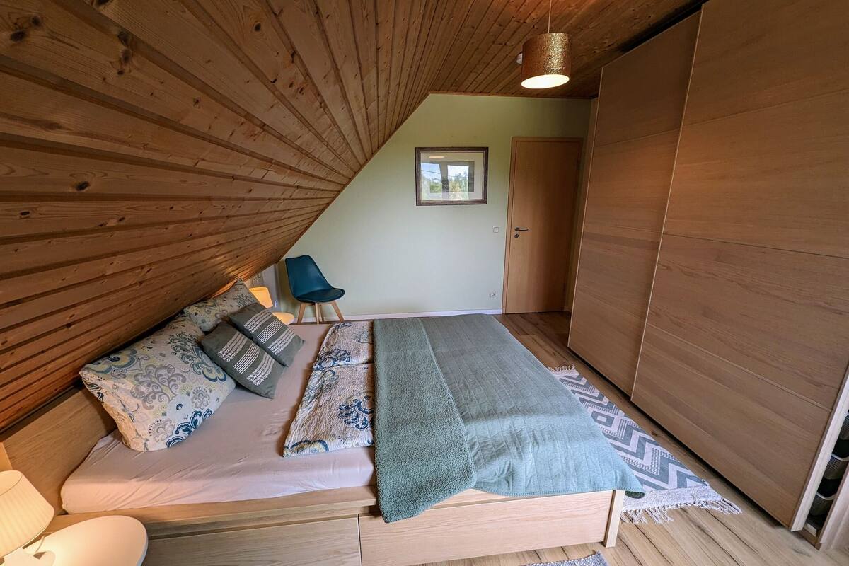A bedroom with a sloped ceiling, a bed with patterned bedding, a blue chair, and a wooden wardrobe.