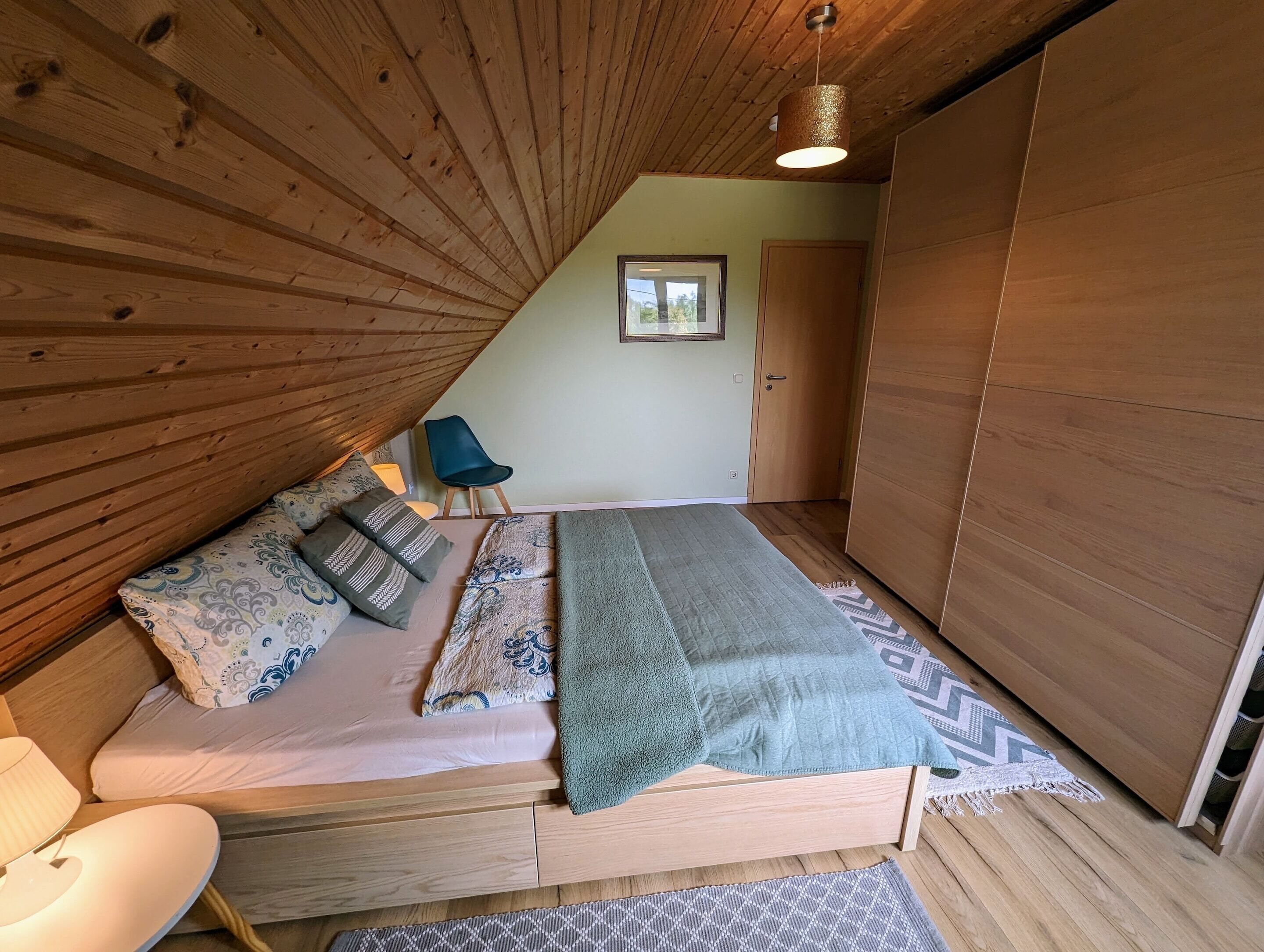 A bedroom with a sloped ceiling, a bed with patterned bedding, a blue chair, and a wooden wardrobe.