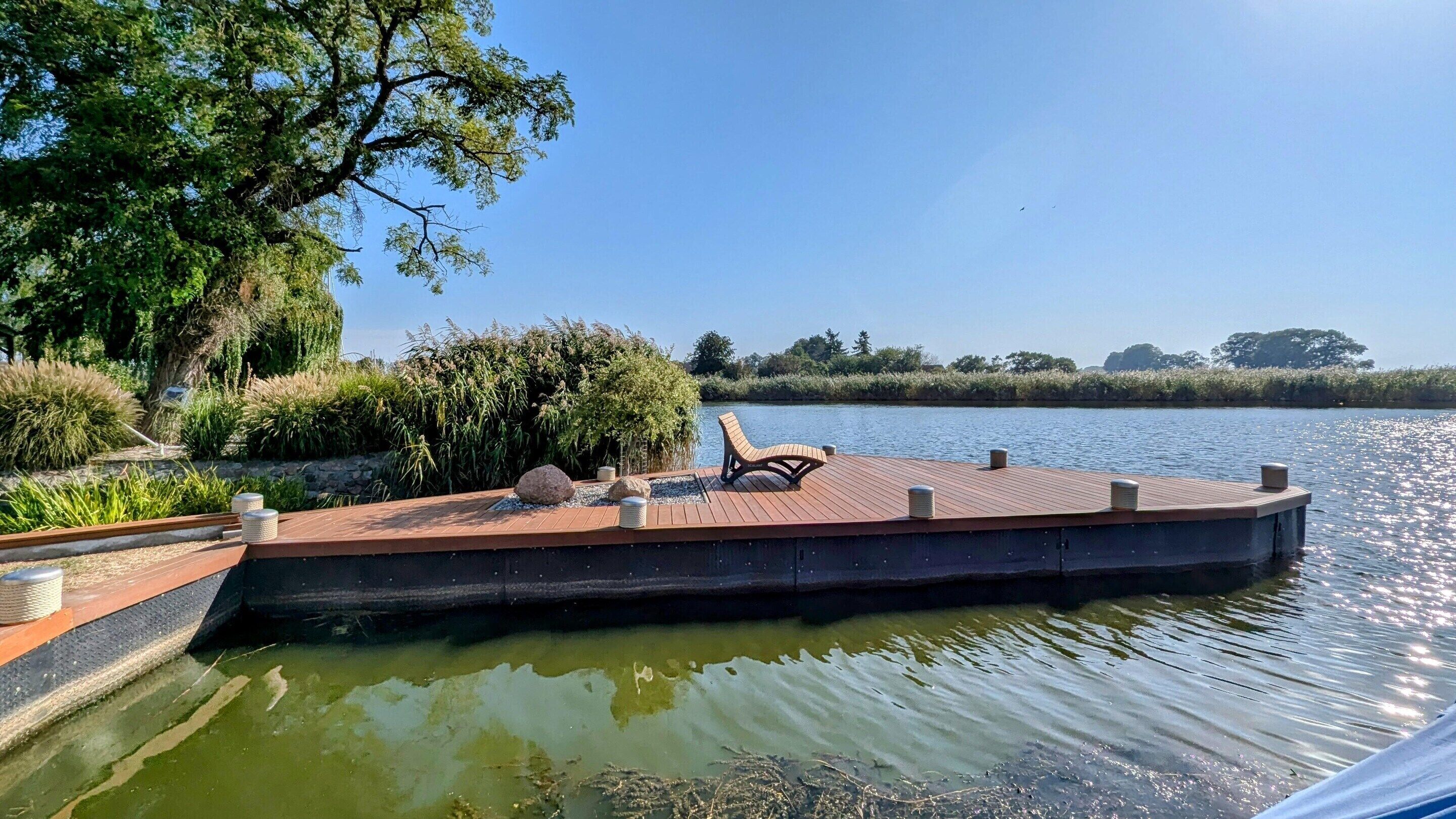 Een houten steiger met een lounge stoel, omringd door groen en kalm water.