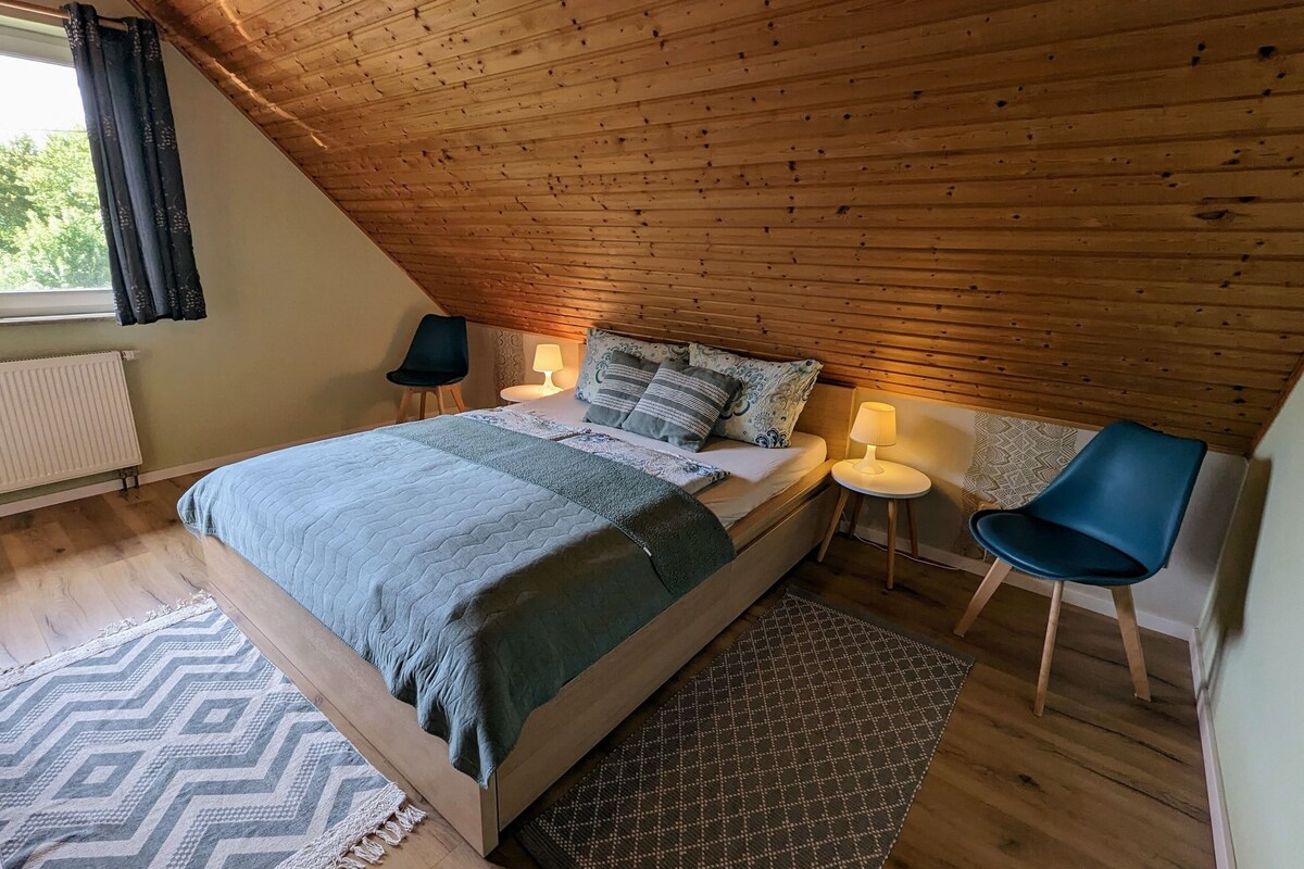 A cozy bedroom with a sloped ceiling, a bed with a patterned bedspread, two bedside lamps, a chair, and a rug.