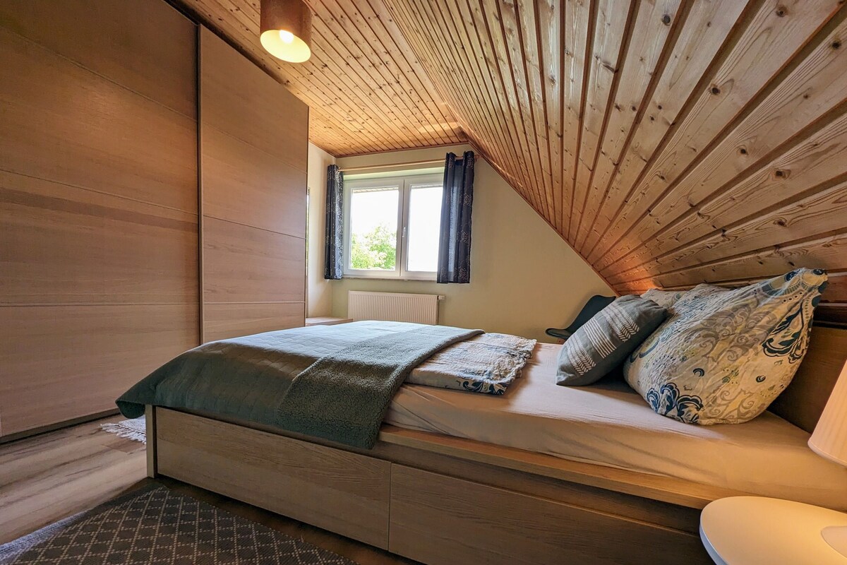 A cozy bedroom with a wooden ceiling, a bed with patterned bedding, a window with curtains, and a radiator.