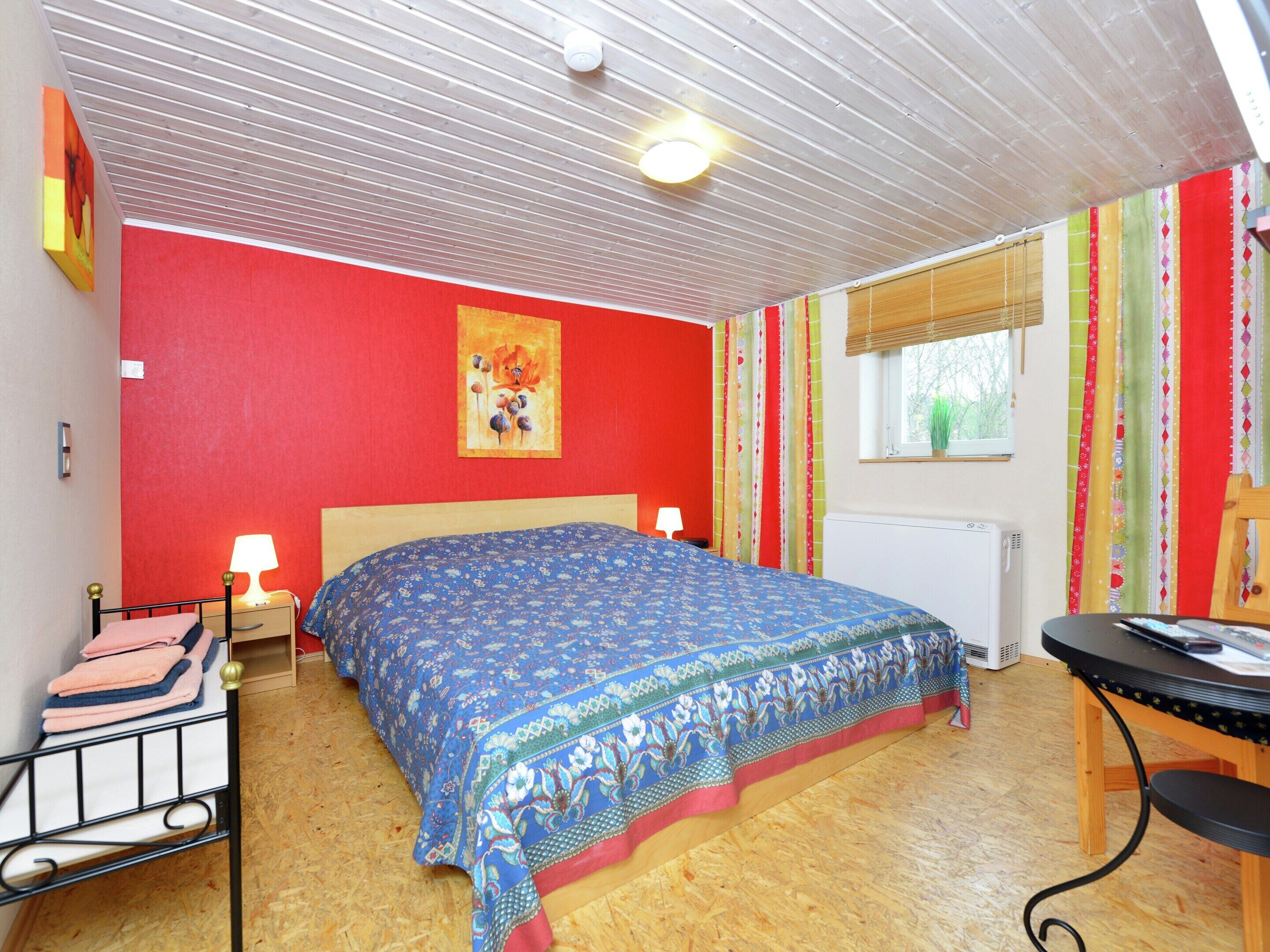 A bedroom with a wooden floor, a bed with a patterned bedspread, a bedside table with a lamp, a radiator, and a window with curtains.