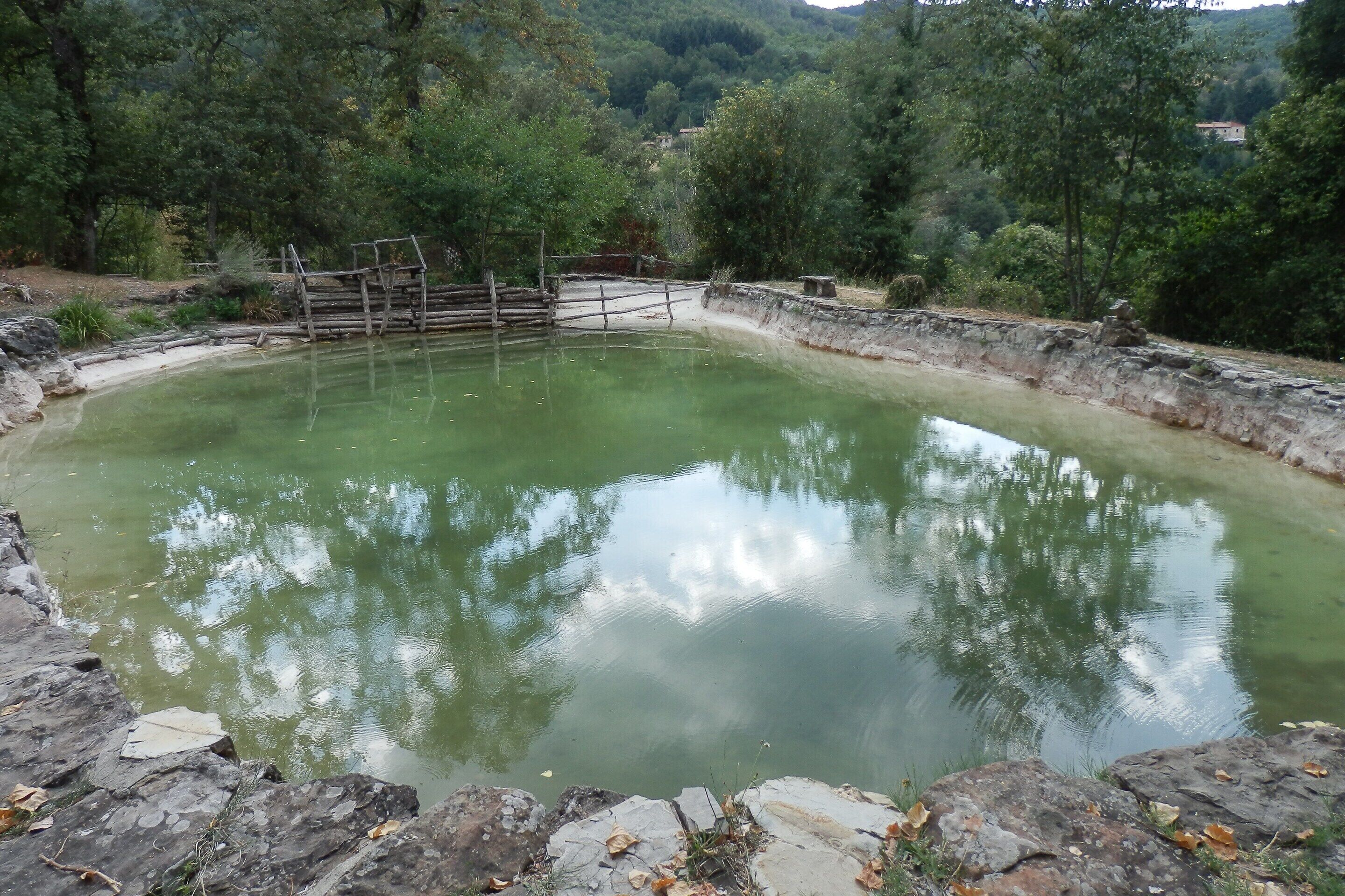 A natural pool with a stone wall and wooden fence, surrounded by trees and hills.