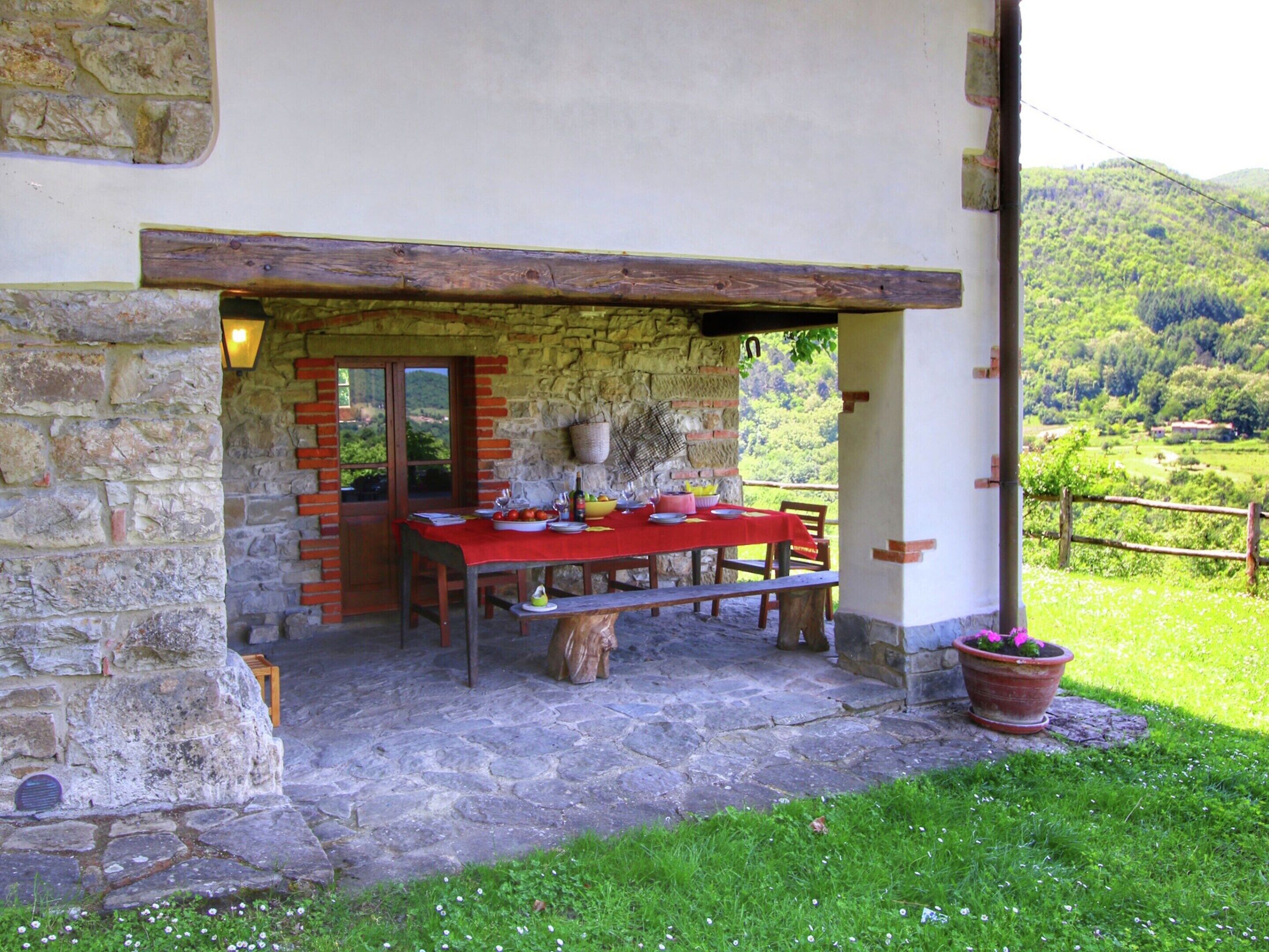 Un bâtiment en pierre avec une table rouge dressée pour le repas, un banc en bois et une plante en pot.