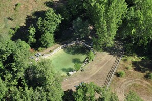 Vista aerea di uno stagno circolare circondato da una fitta foresta, con un sentiero sterrato che conduce a un ponte di pietra.