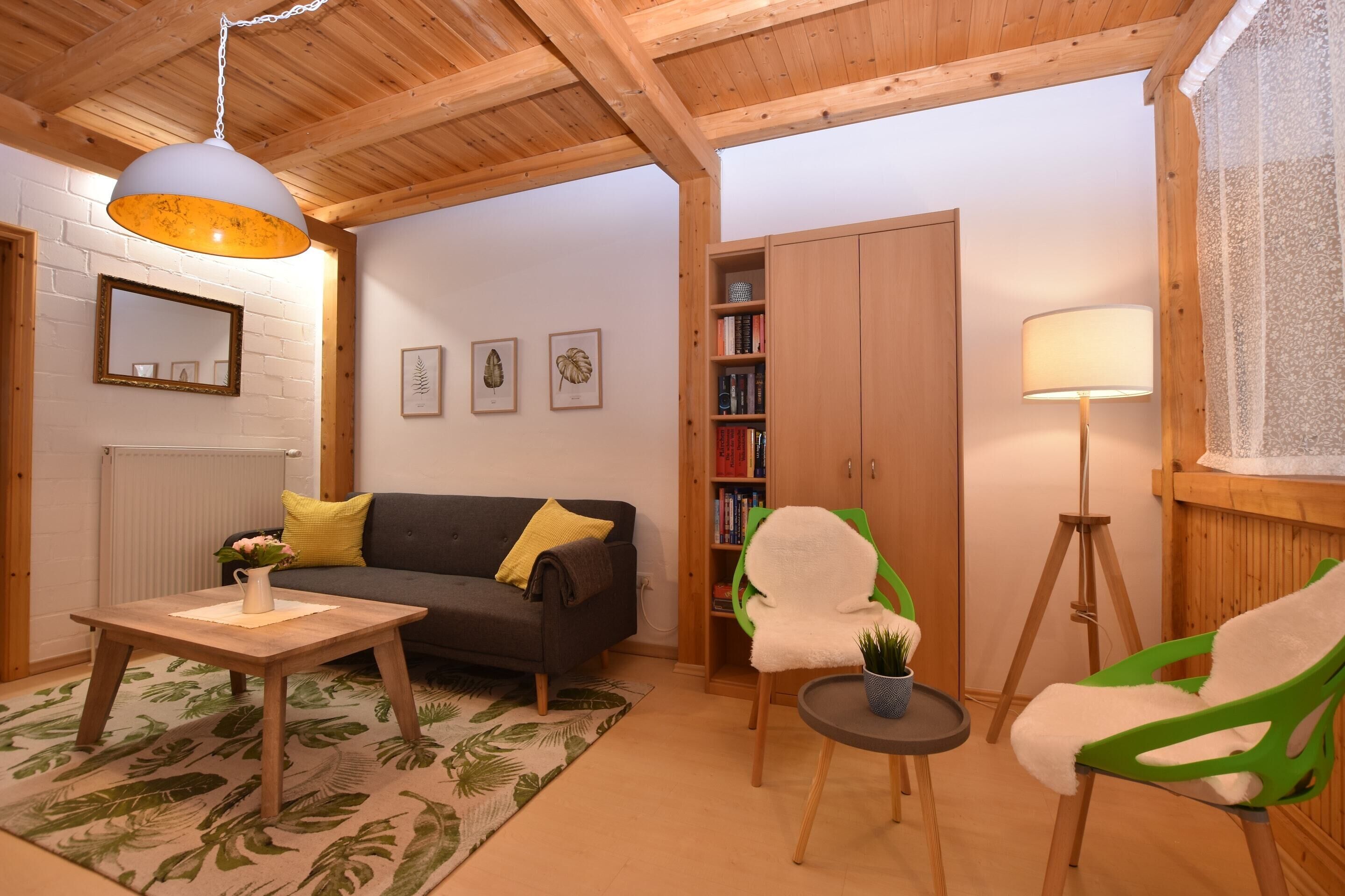 Ein gemütliches Wohnzimmer mit Holzbalkendecke, einem grauen Sofa, einem kleinen Tisch mit einer Vase, einem grünen Sessel und einem Bücherregal.