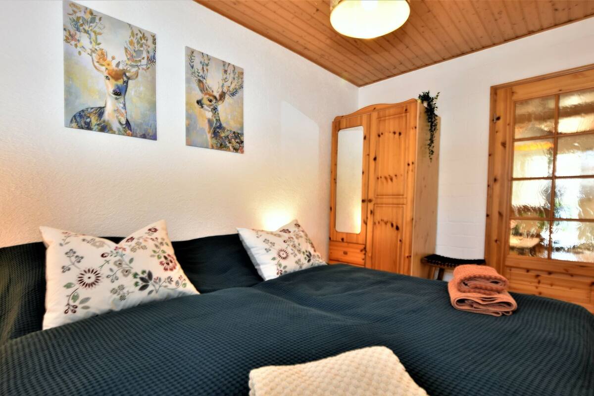 A bedroom with a bed, pillows, a wooden wardrobe, and two paintings on the wall.