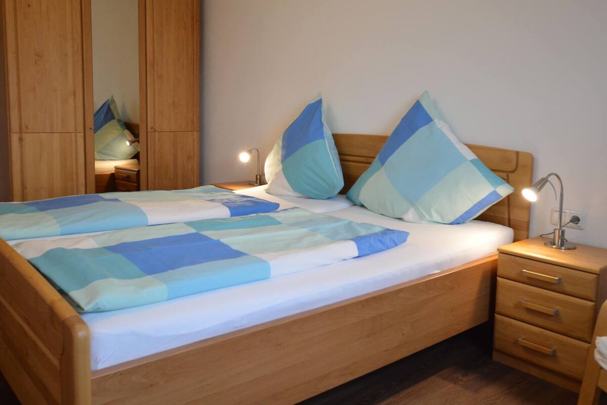 A neatly made bed with a blue and white checkered bedspread and two large pillows. There is a wooden wardrobe to the left and a bedside table with a lamp on the right.