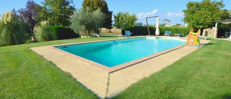 Una piscina rectangular con borde de azulejos, rodeada de césped y árboles bien cuidados.