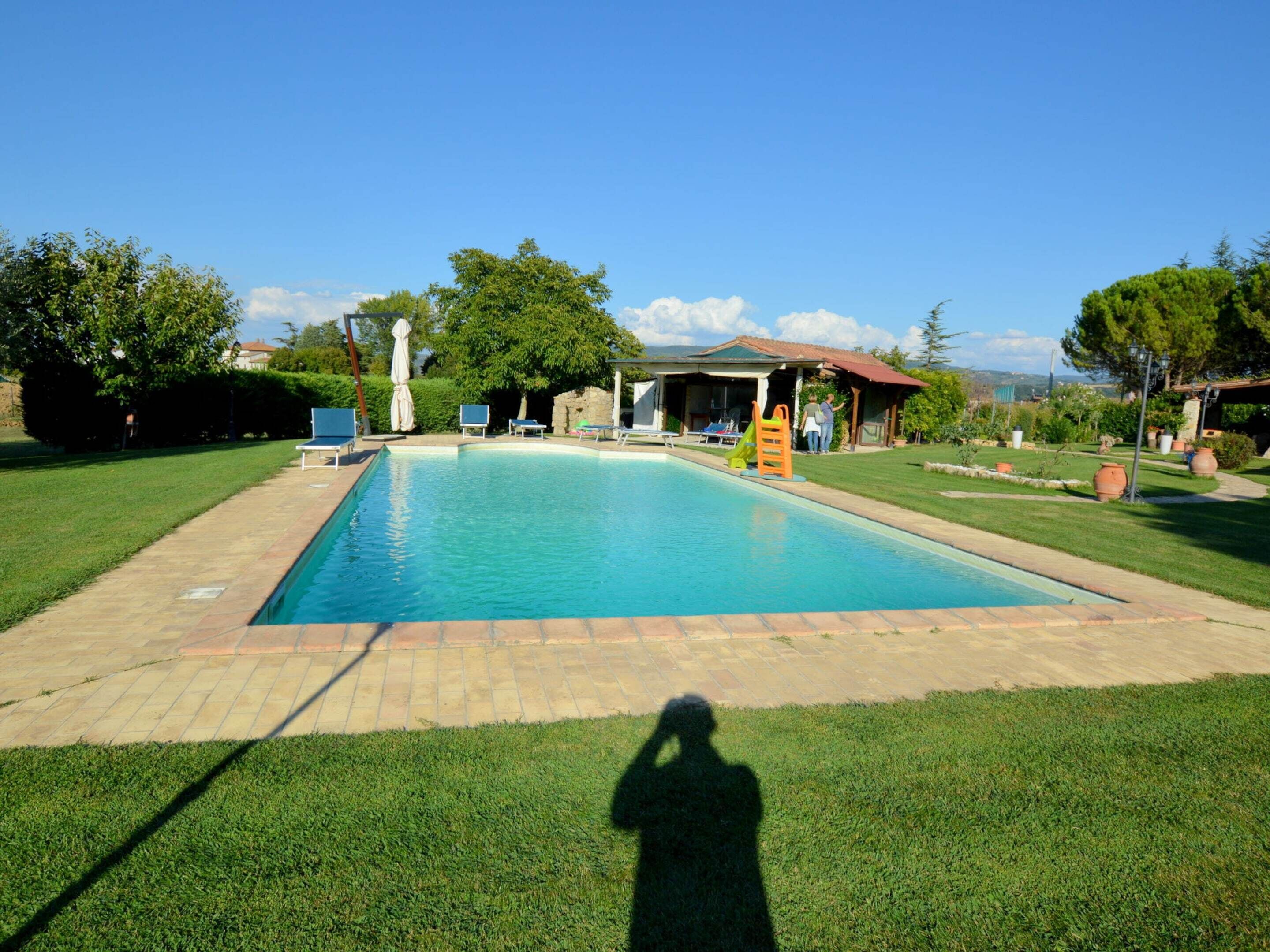 A rectangular swimming pool with a tiled deck, surrounded by well-maintained grass, a covered seating area, and a small play structure.