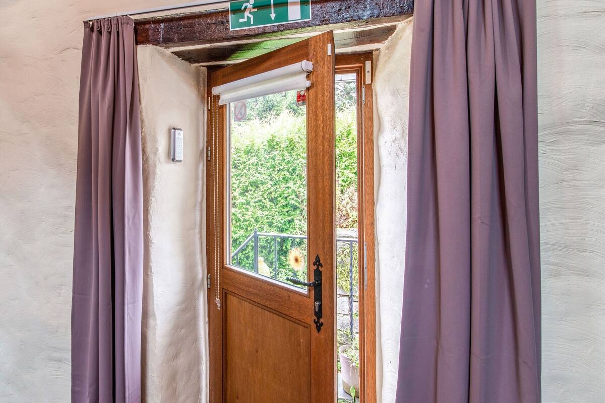 Une porte en bois surmontée d’un panneau vert indiquant la sortie de secours, une fenêtre donnant sur un paysage verdoyant et des rideaux violets de chaque côté.
