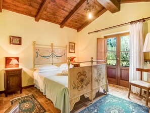 Un dormitorio con techo de madera, una cama con cabecera decorativa, una mesita de noche, un ventanal con cortinas y vistas al exterior con vegetación.