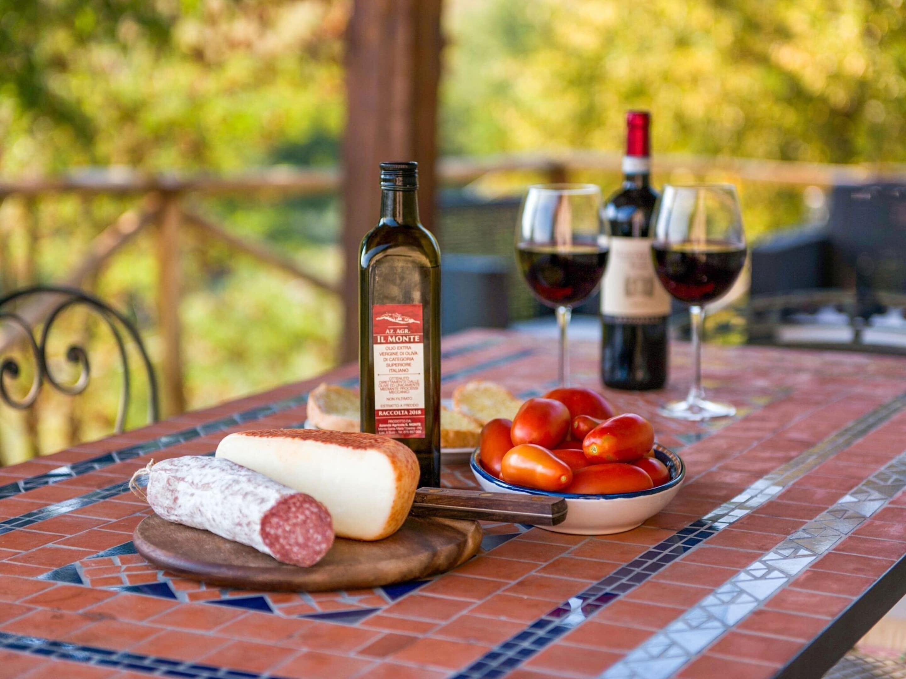 A table with a bottle of wine, two glasses of wine, a bowl of tomatoes, and a plate with a cheese and a cured meat.
