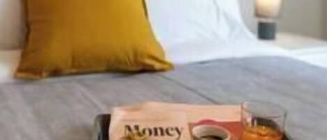 A neatly made bed with a white pillow and a mustard-colored pillow. A tray with a newspaper, a cup, and a glass of orange juice is placed on the bed.