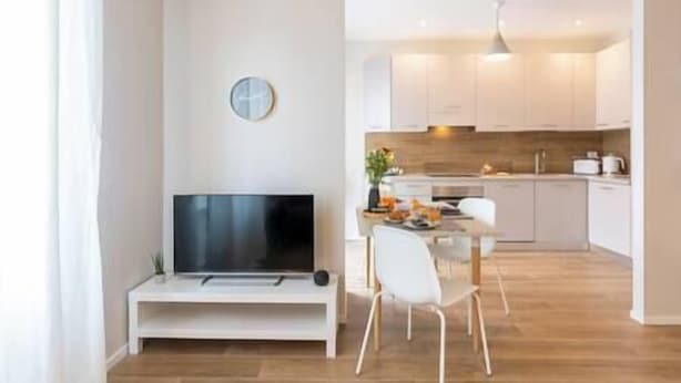 A modern living room with a flat-screen TV, a small white table, and a dining area with a white chair.
