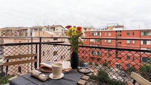A balcony with a table set for two, a vase of flowers, and a view of surrounding buildings.