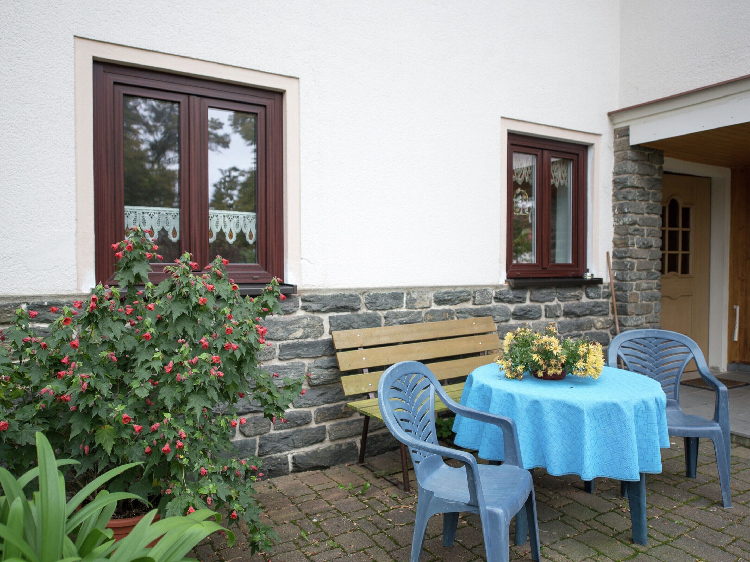 Une terrasse avec une table et des chaises, un banc et un pot de fleurs orné de fleurs rouges.
