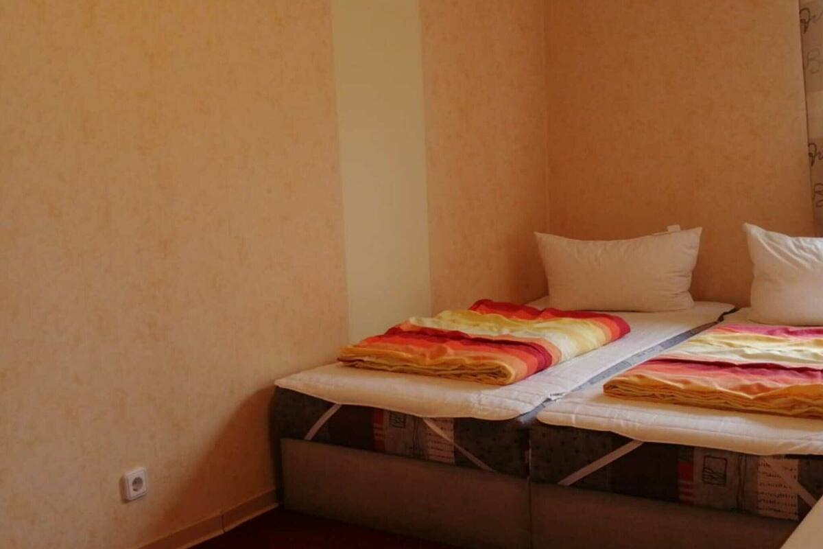 A small, single-bed room with a mattress on the floor, a wall socket, and plain walls.