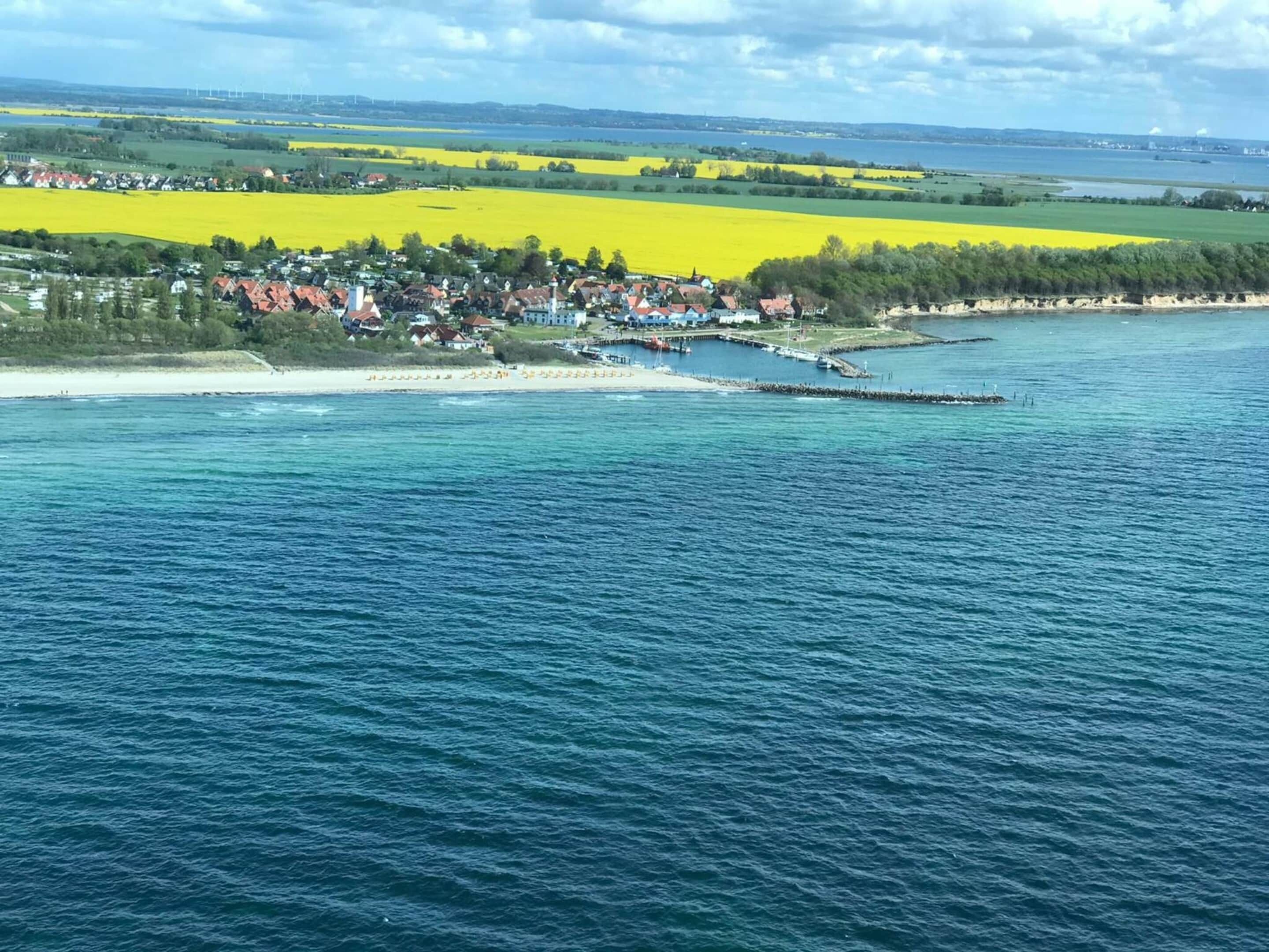Rannikkokaupunki, jossa on laituri, joka ulottuu merelle, ja jota ympäröivät keltaisten kukkien niityt. Taustalla näkyy tuulivoimaloita.
