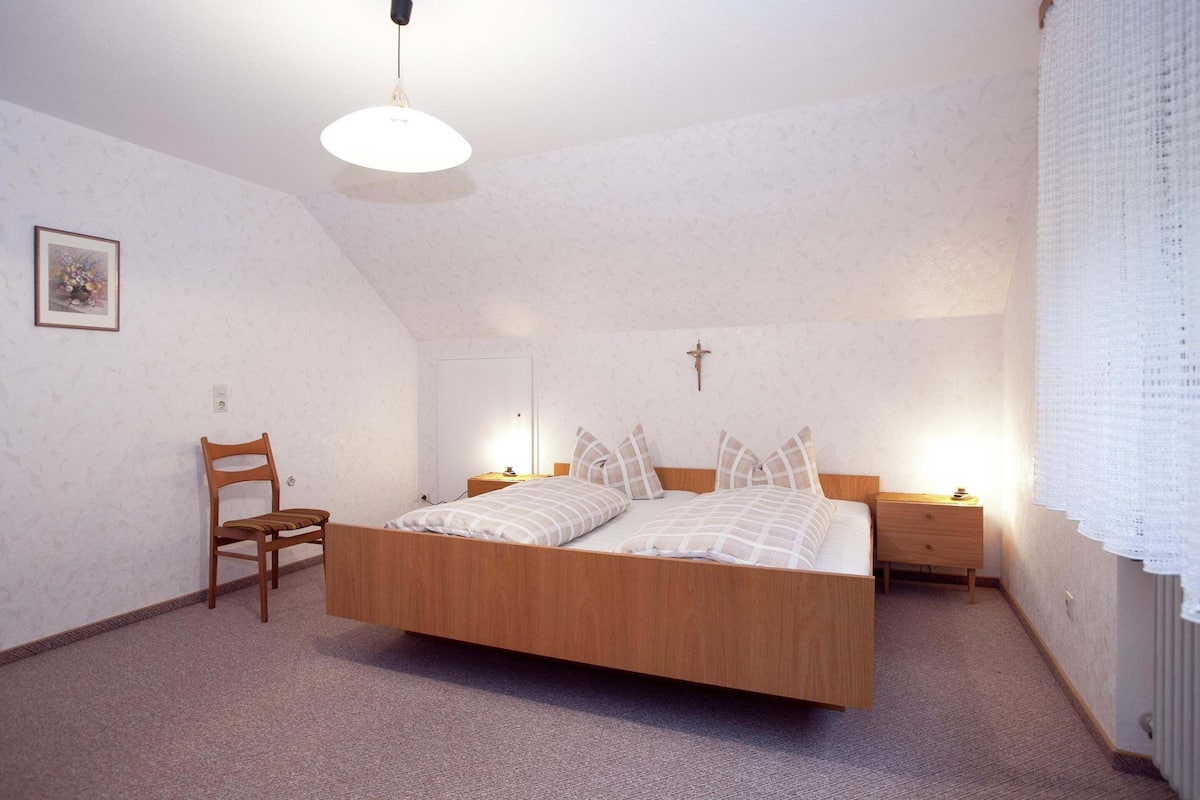 A bedroom with a wooden bed, a chair, a nightstand, and a wall-mounted light fixture.