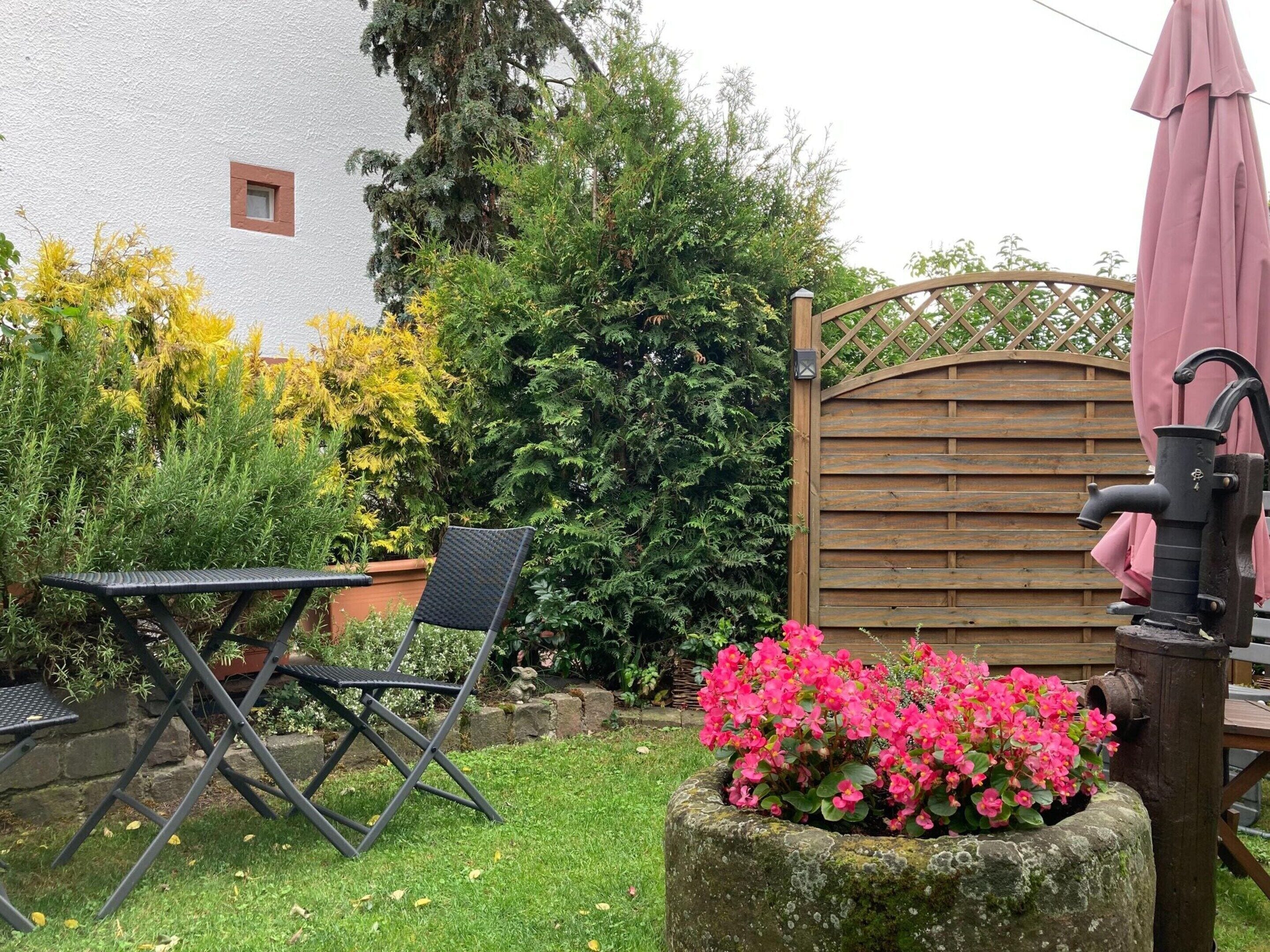 A garden with a wooden gate, a metal table and chair, and a flower pot with pink flowers.