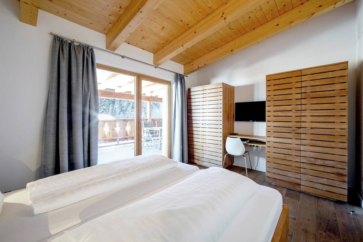 A bedroom with a wooden ceiling, a bed with white bedding, a desk with a chair, and a TV on a wooden cabinet.