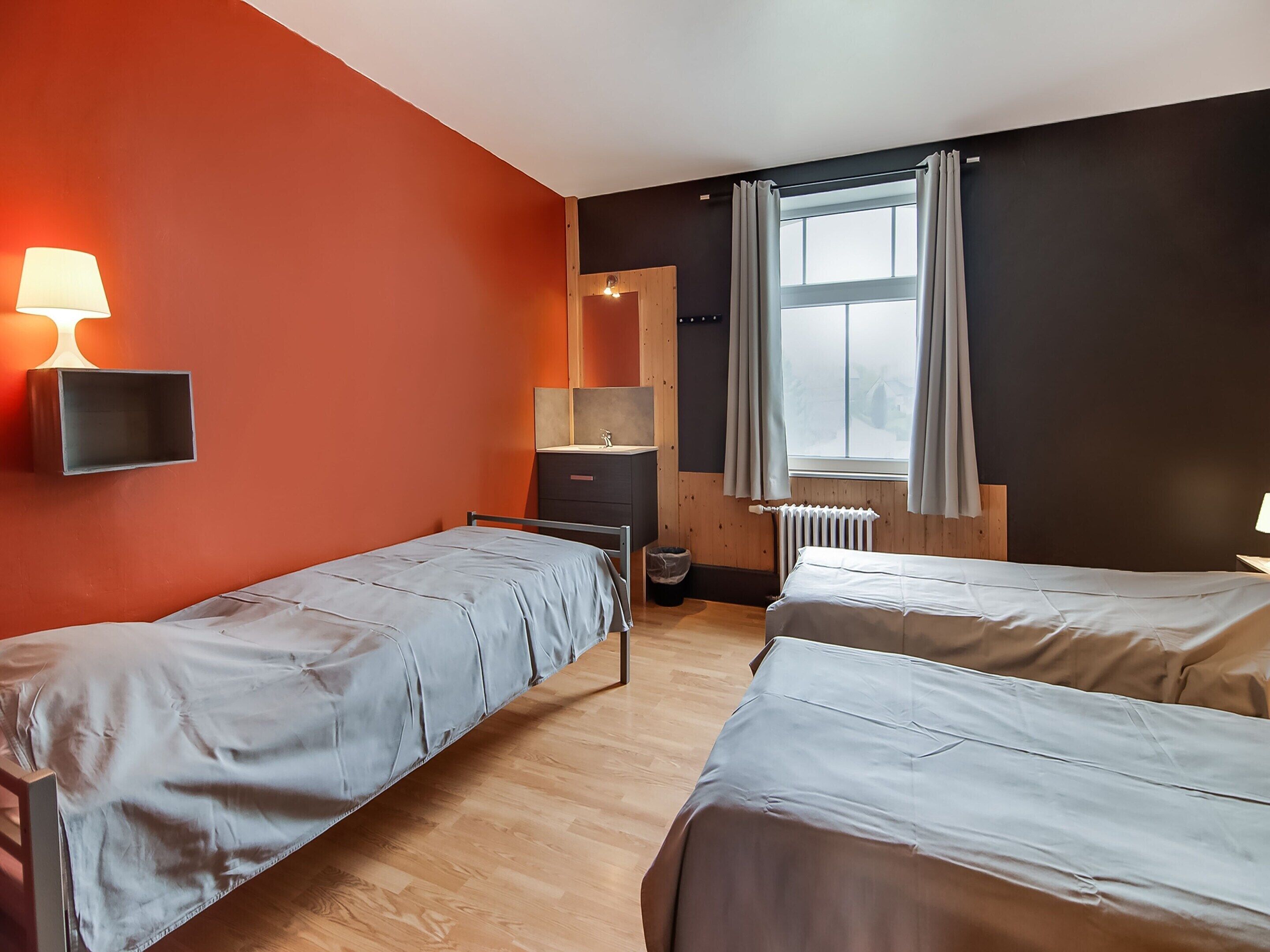 A room with two single beds, a wooden dresser, a mirror, a radiator, and a window with curtains.