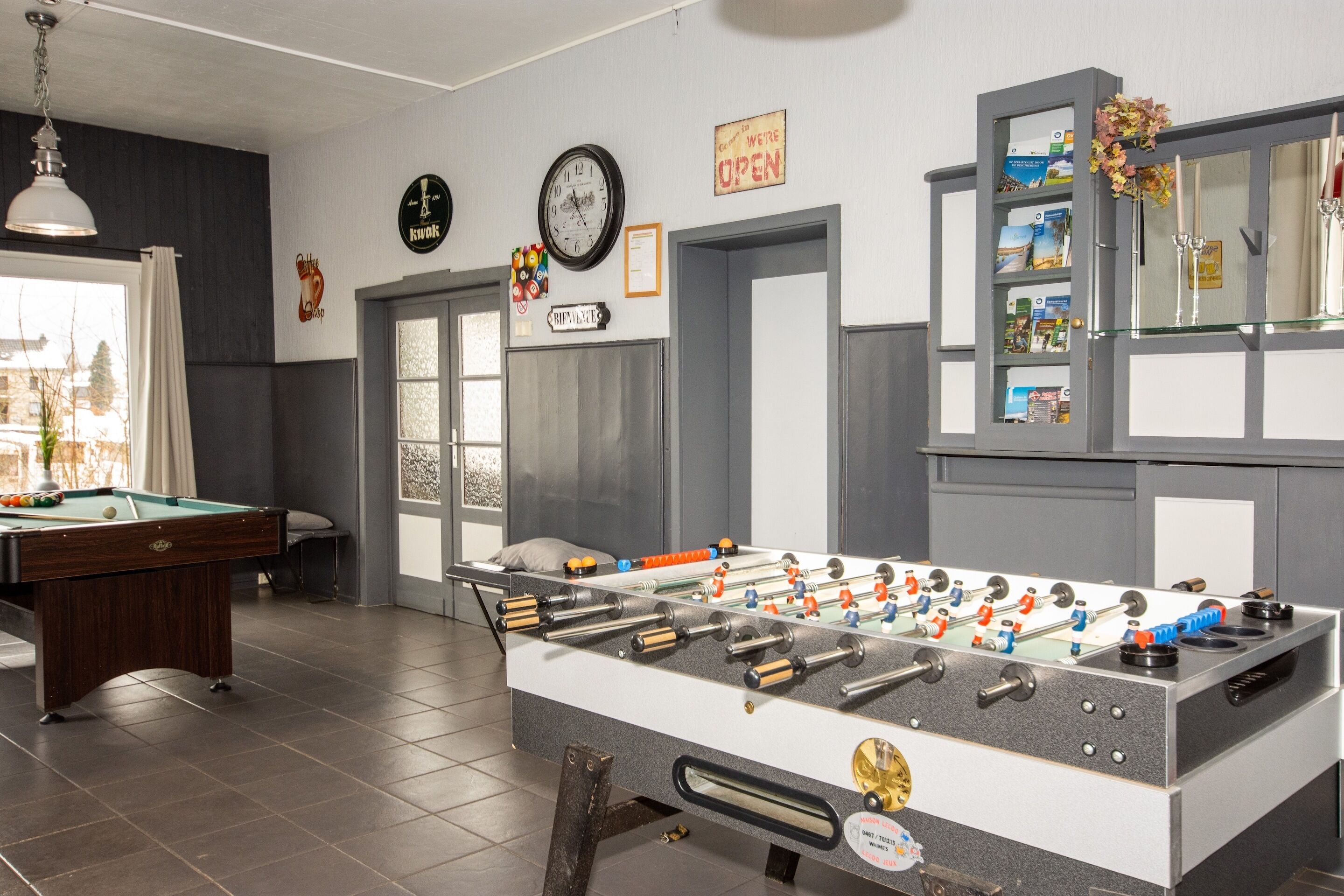 A room with a foosball table, a pool table, and a billiards table. There are doors, a clock, and a sign that says "We're Open".