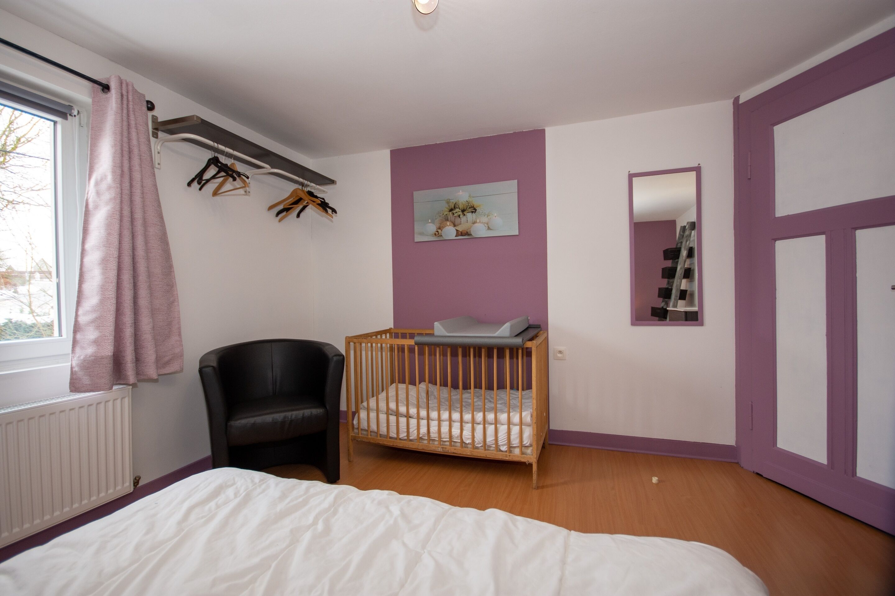 Ein Schlafzimmer mit einem Babybett, einem Sessel, einer Heizung und einem Fenster mit Vorhang.
