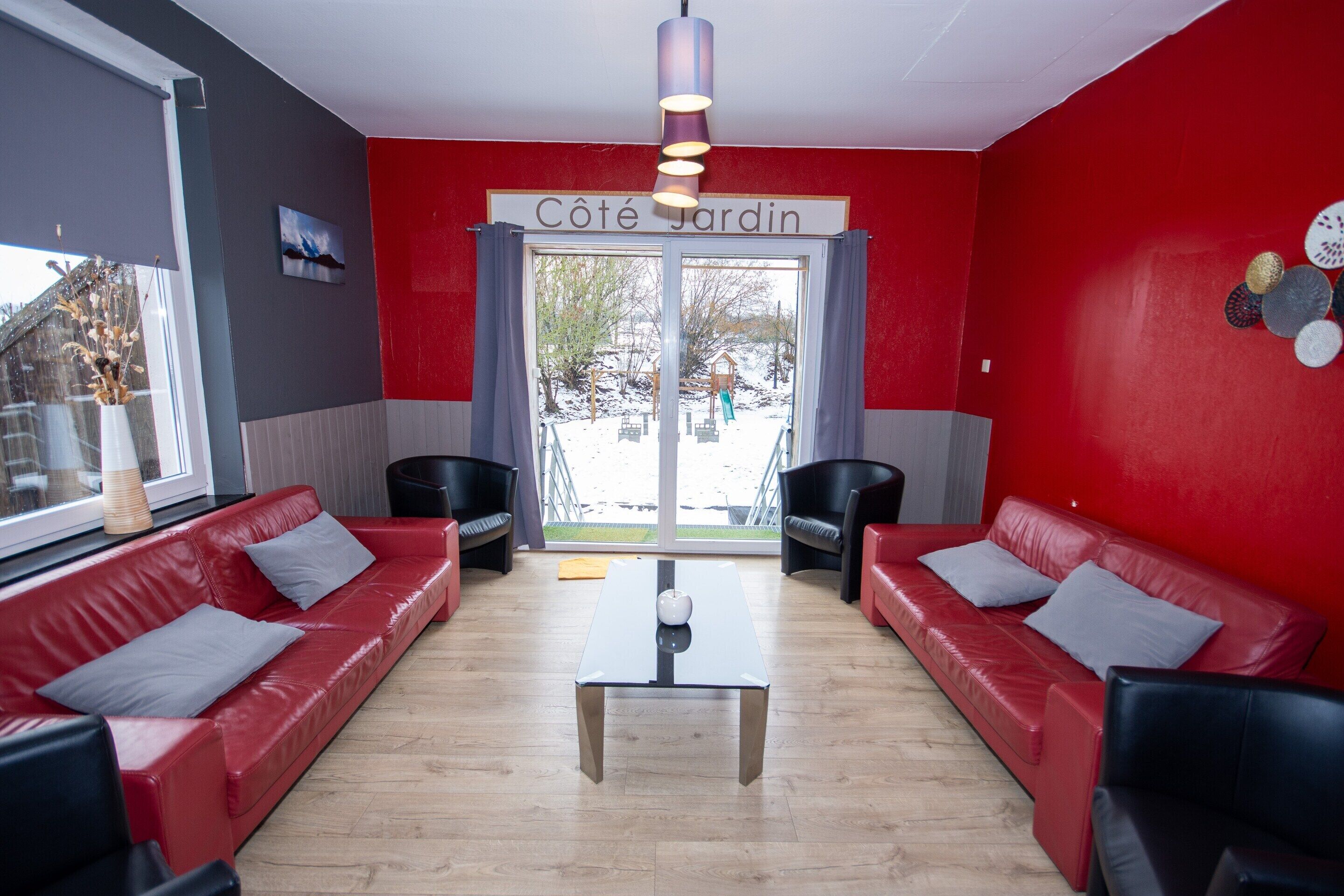 Una habitación en tonos rojo y gris con dos sofás rojos, una mesa de centro y un ventanal con vistas a un parque infantil.