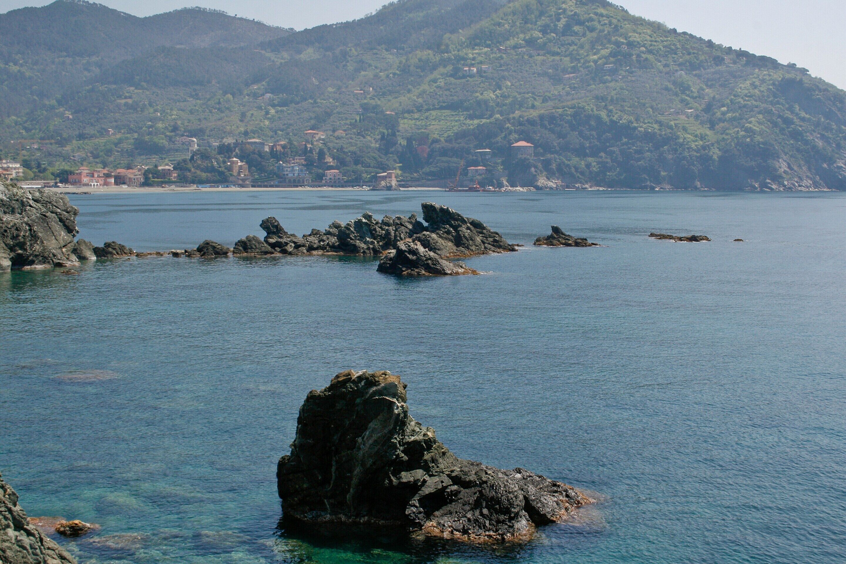 A coastal landscape with rocky outcrops in the water and a hilly background with buildings.