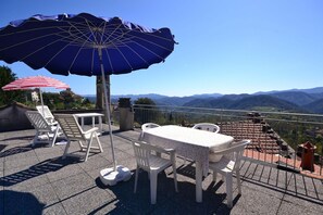 Terraza exterior con mesa y sillas, una sombrilla azul y vistas a montañas.