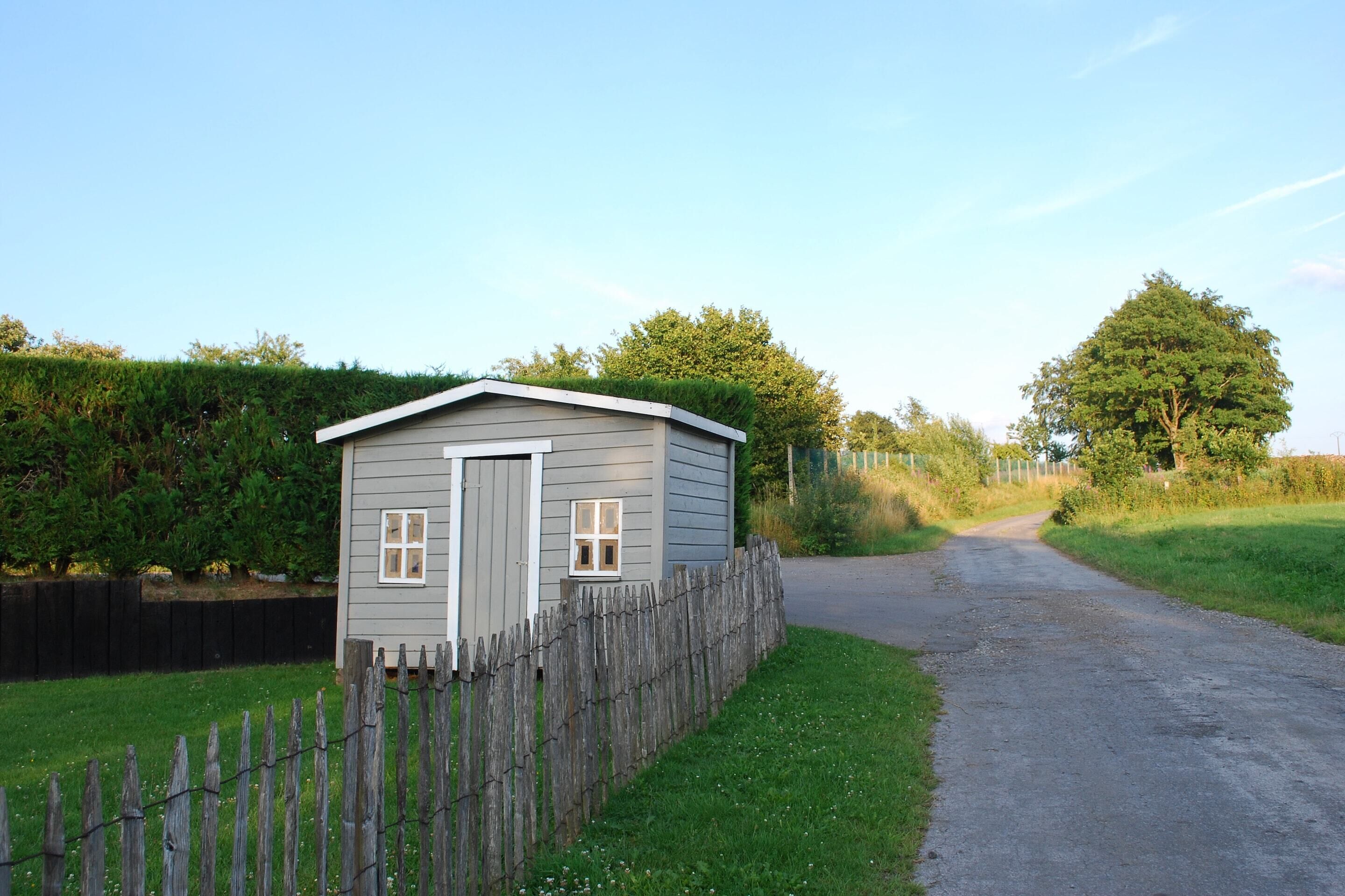 A small, single-story shed with a door and two windows, surrounded by a wooden fence and a gravel path leading to a road.