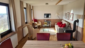 A modern living room with a wooden floor, beige sofas, a dining table with fruit, and a TV mounted on the wall.