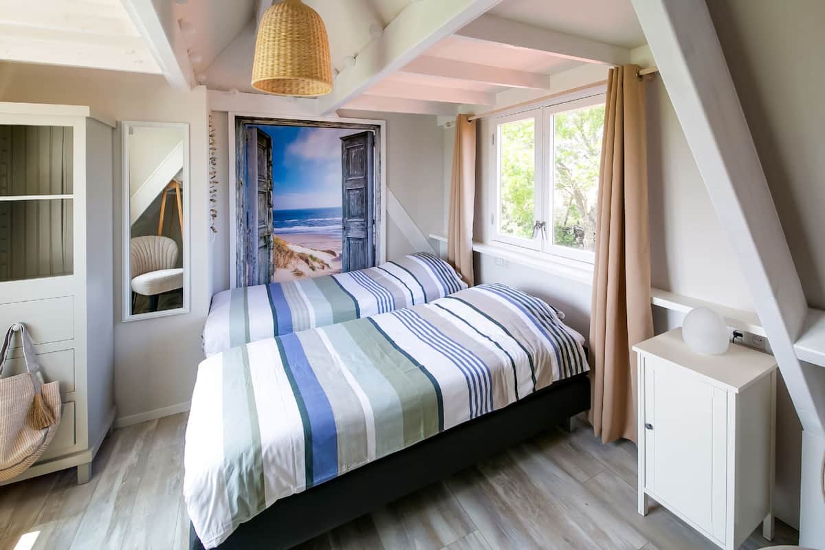 A bedroom with a bed, a wardrobe, a hanging light fixture, a window with curtains, and a view of the beach through an open door.