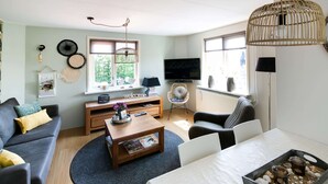 A living room with a sofa, coffee table, and TV. There is a window with blinds, a lamp, and a decorative item on the wall. - Beautiful Apartment in Callantsoog Near Beach (t Zand)