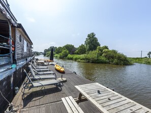 A wooden deck with lounge chairs and a kayak, overlooking a calm river with greenery and trees. - Cushy Boat in Merkem With Terrace (Houthulst)