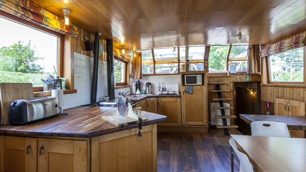 A wooden cabin-style kitchen with a wooden countertop, a toaster, a kettle, a microwave, and a sink. There are windows with curtains, a wooden ceiling, and a view of greenery outside. - Cushy Boat in Merkem With Terrace (Houthulst)