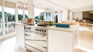 A modern kitchen with a built-in oven, a dining area with a table and chairs, and large windows offering a view of the outdoors. - Luxurious Villa With Pool and Sauna, and Very Suitable for Culinary Workshops! (Waimes)