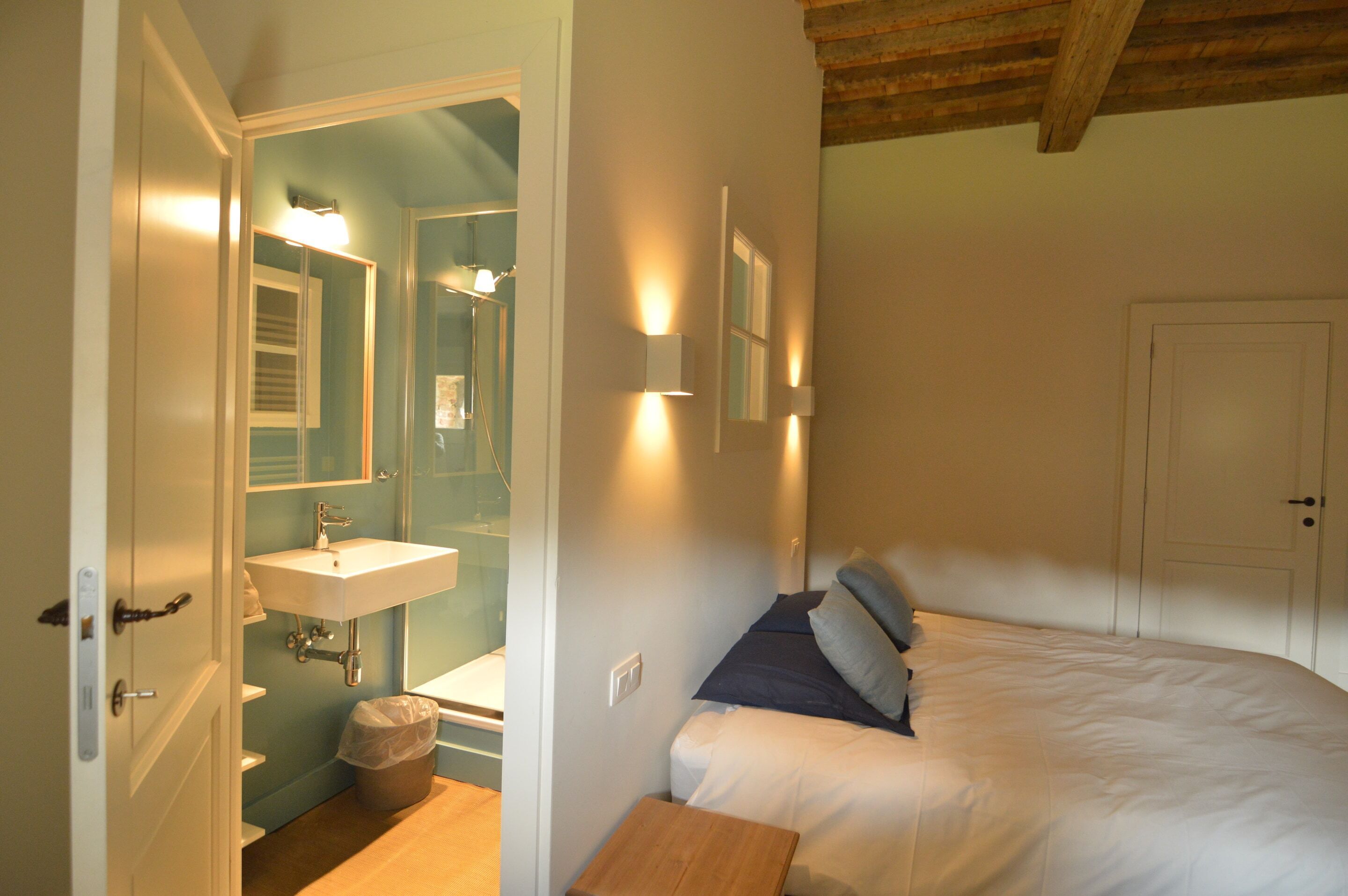 A bedroom with a bed, a bedside table, a nightstand, a wall-mounted light, a mirror, a window, and a door.