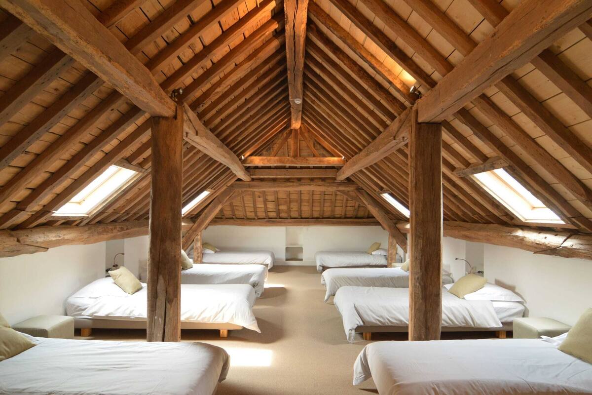 A room with four beds arranged in a U-shape, wooden beams on the ceiling, and skylights.