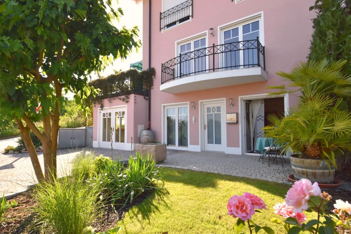 Un edificio rosa con un balcone, un giardino con erba e piante e una porta con una finestra.