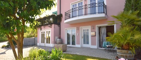 A pink building with a balcony, a garden with grass and plants, and a door with a window.