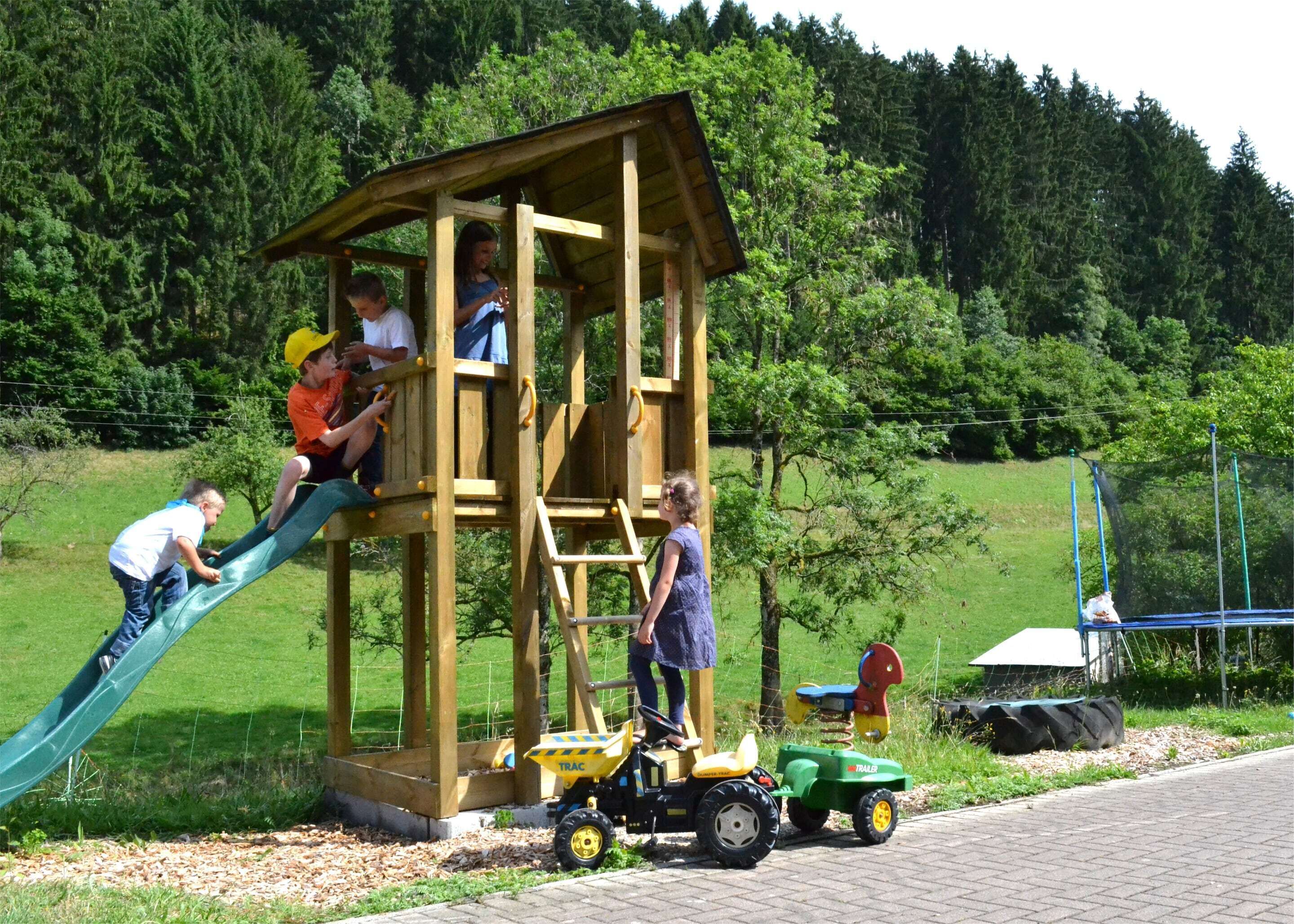 Une structure de jeux en bois, dotée d’un toboggan et d’une zone d’escalade, entourée d’un champ herbeux et d’arbres.