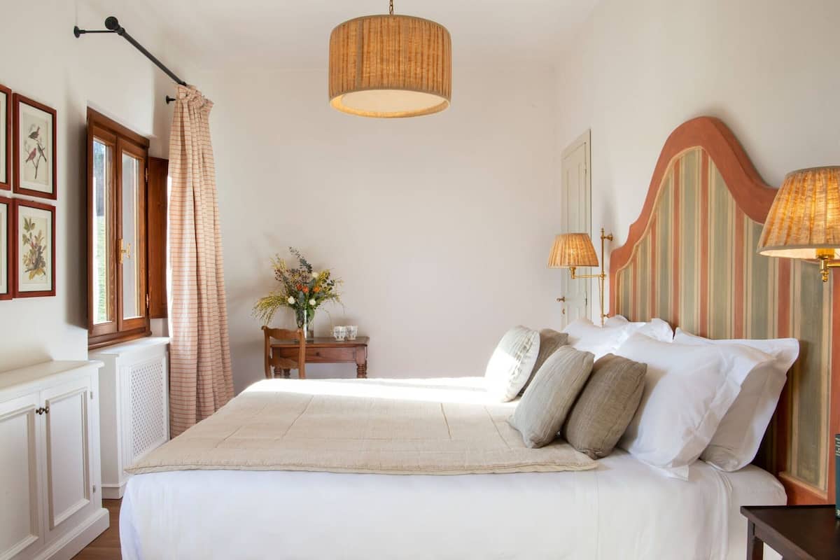 Une chambre avec un grand lit, une tête de lit en bois, un luminaire suspendu, une table de chevet avec une lampe et une fenêtre avec des rideaux.