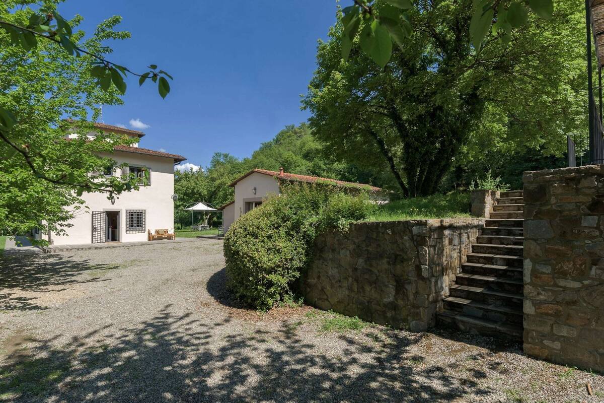 Une propriété rurale avec une allée de gravier menant à un bâtiment blanc doté d’un toit en tuiles, entouré d’arbres et d’un mur de pierre avec un escalier.