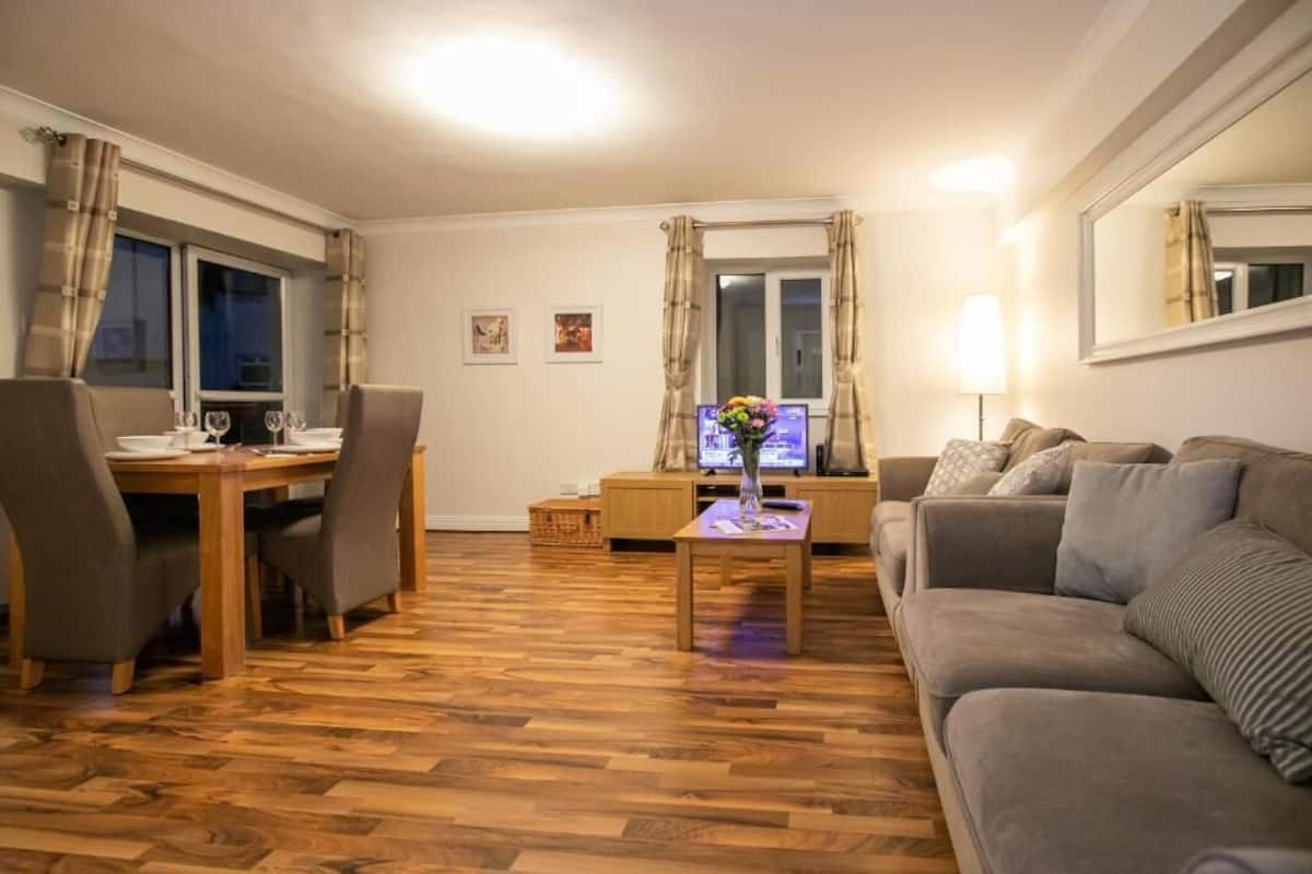 Una sala de estar con piso de madera, una mesa de comedor con sillas, un sofá, un televisor y una lámpara.